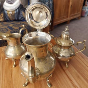 Op de afbeelding: Een driedelige theeset van messing, bestaande uit een theepot met een open scharnierend deksel, een melkkan en een suikerpot met deksel. Elk item is voorzien van een gegraveerd bloemmotief, gebogen handvatten en drie kleine pootjes.