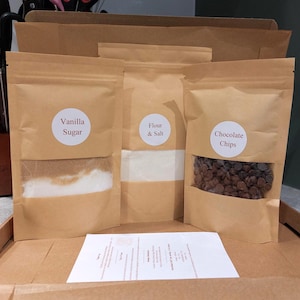 May include: A baking kit featuring three brown paper pouches. The pouches are labelled "Vanilla Sugar", "Flour & Salt", and "Chocolate Chips". The contents are visible through the transparent sections. A white paper with text is in front.