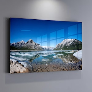 May include: A landscape photograph of a mountain range reflected in a lake. The sky is a clear, bright blue. The mountains have snow-capped peaks and green slopes. The lake has patches of ice and clear water, with rocks visible at the bottom.