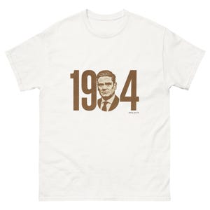 May include: White t-shirt featuring a brown graphic of the number "1984" with a portrait of a man inside the "0". The text "defec.world" is printed below the graphic. The shirt has short sleeves and a crew neck.