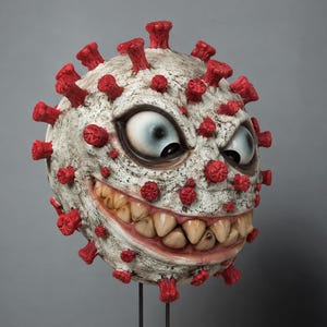 May include: A spherical sculpture resembling a virus with a textured off-white surface and numerous protruding red spikes. The object features a wide, menacing grin with realistic human-like teeth and two large, wide-eyed, expressive cartoon eyes.