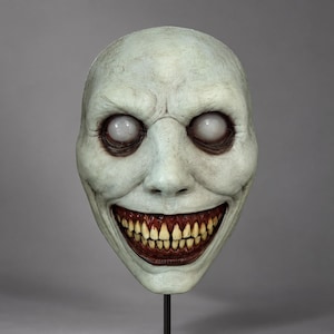 May include: A pale, off-white full-head mask featuring a wide, toothy grin with yellowed, realistic teeth and dark, recessed gums. The mask has smooth, featureless white eyes and deep, dark shadows around the eye sockets and mouth area.