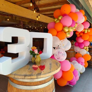 Könnte beinhalten: Ein Holztisch in Form eines Fasses mit der weißen Zahl "30", umgeben von einem Ballonbogen in Rosa, Orange und Gold. Zwei kleine Vasen mit Rosen und herzförmigen Sonnenbrillen stehen auf dem Tisch. Die Szene spielt sich im Freien ab.