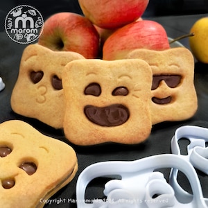 Peut inclure: Gros plan sur plusieurs biscuits carrés avec divers visages d'emoji en chocolat. Les biscuits sont brun clair avec des garnitures au chocolat noir. Des pommes rouges sont en arrière-plan. Des emporte-pièces blancs sont au premier plan. Le texte "Copyright Marommolds 2026. All rights reserved" est en bas.
