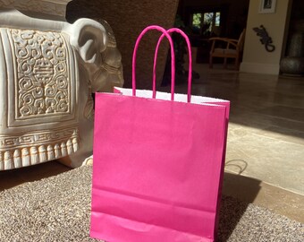 May include: A bright pink paper gift bag with matching pink handles stands on a textured beige rug. The bag is empty and has a white interior. The background includes a decorative white stone sculpture and a glimpse of a living room.