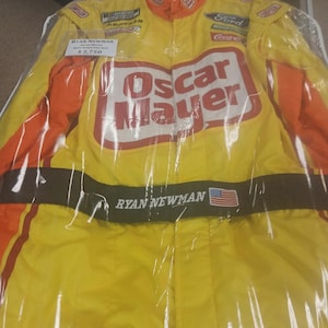 Puede incluir: Un mono de carreras amarillo y naranja con el logo "Oscar Mayer". El mono presenta el nombre "Ryan Newman" y la bandera estadounidense. Está dentro de una bolsa de plástico transparente.