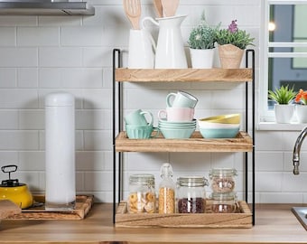 May include: A three-tiered wooden shelf with black metal supports, holding various kitchen items. The top shelf displays white pitchers, utensils, and potted plants. The middle shelf has pastel-colored bowls and cups. The bottom shelf features glass jars with food items.