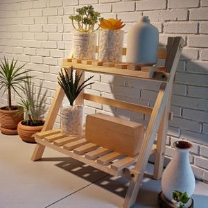 Puede incluir: Un estante de madera de dos niveles para plantas con acabado natural. El estante contiene varias plantas en macetas y objetos decorativos, incluyendo un jarrón de cerámica blanca, un bloque de madera y jarrones de vidrio llenos de piedras blancas y vegetación. El fondo es una pared de ladrillo blanco.