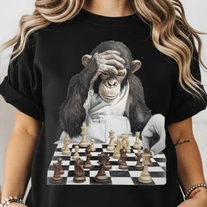 May include: Black crew neck t-shirt featuring a detailed illustration of a chimpanzee with its hand on its forehead, seemingly perplexed while playing chess. The chess board has black and white squares and chess pieces.