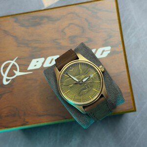 May include: A gold-toned wristwatch with a brown leather strap sits on a grey cushion. The watch face has an embossed plane design. The watch is displayed on a wooden box with the Boeing logo.