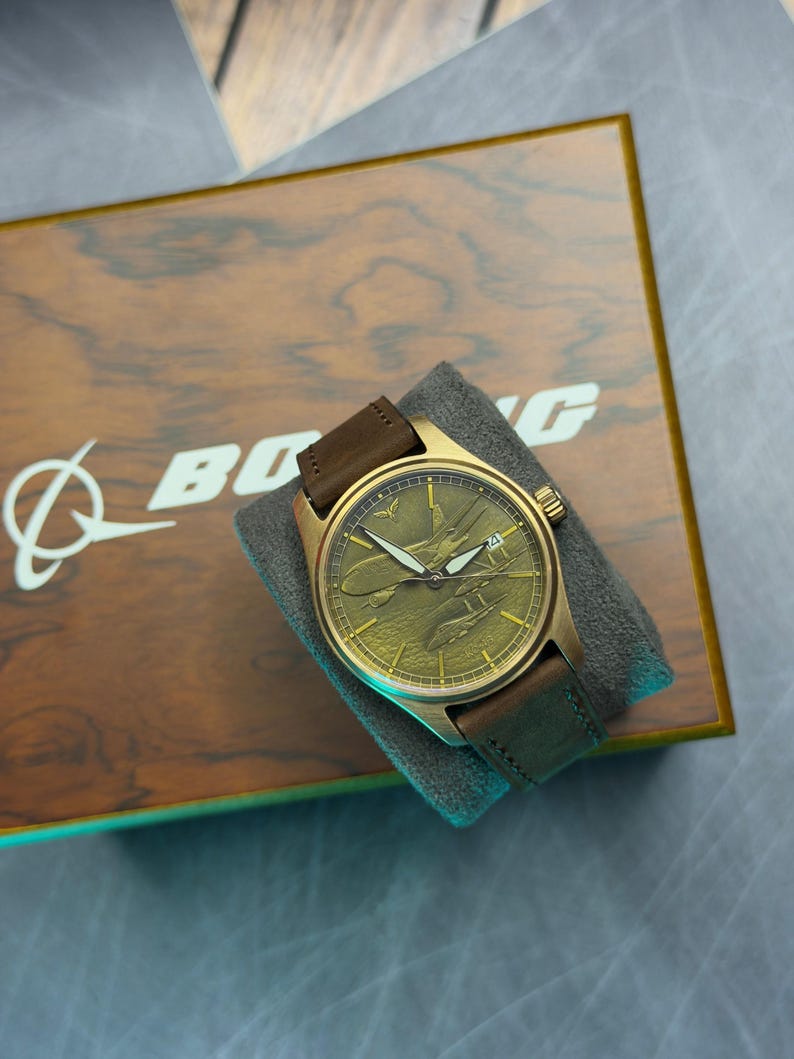 May include: A gold-toned wristwatch with a brown leather strap sits on a grey cushion. The watch face has an embossed plane design. The watch is displayed on a wooden box with the Boeing logo.