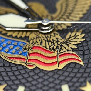 K&ouml;nnte beinhalten: Nahaufnahme eines Zifferblatts mit einem goldenen Adler, der eine Flagge mit roten und wei&szlig;en Streifen und blauen Sternen h&auml;lt. Das Zifferblatt ist dunkelgrau mit goldenen Sternstundenmarkierungen und wei&szlig;en Zeigern.