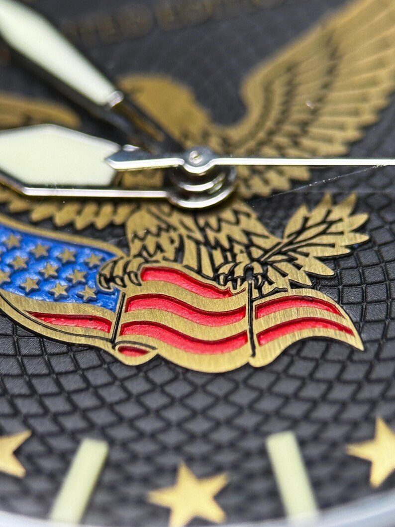 K&ouml;nnte beinhalten: Nahaufnahme eines Zifferblatts mit einem goldenen Adler, der eine Flagge mit roten und wei&szlig;en Streifen und blauen Sternen h&auml;lt. Das Zifferblatt ist dunkelgrau mit goldenen Sternstundenmarkierungen und wei&szlig;en Zeigern.