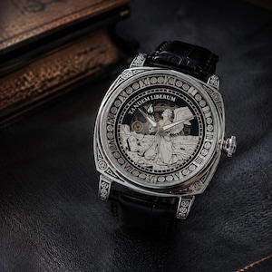 Puede incluir: Un reloj de pulsera plateado con correa de cuero negro y caja cuadrada. La esfera presenta una ilustraci&oacute;n grabada de un &aacute;ngel arrodillado en un cementerio, el texto TANDEM LIBERUM y n&uacute;meros romanos a lo largo del borde exterior del reloj.