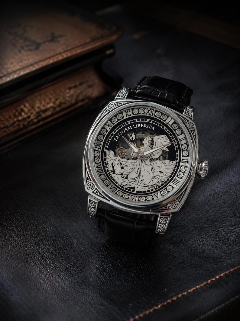 Puede incluir: Un reloj de pulsera plateado con correa de cuero negro y caja cuadrada. La esfera presenta una ilustraci&oacute;n grabada de un &aacute;ngel arrodillado en un cementerio, el texto TANDEM LIBERUM y n&uacute;meros romanos a lo largo del borde exterior del reloj.