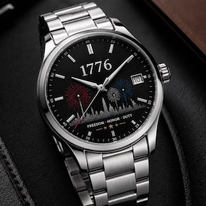 以下が含まれることがあります： 黒い文字盤のステンレススチール製腕時計。 文字盤には「1776」の数字、花火、そして「FREEDOM HONOR DUTY」の文字が描かれています。 時計には日付表示があり、シルバーのバンドが付いています。