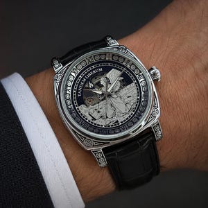 Puede incluir: Un reloj de pulsera plateado con correa de cuero negro, que presenta una caja cuadrada con grabados intrincados. La esfera muestra una ilustraci&oacute;n central de un &aacute;ngel, el texto TANDEM LIBERUM y un borde con n&uacute;meros romanos y motivos circulares.
