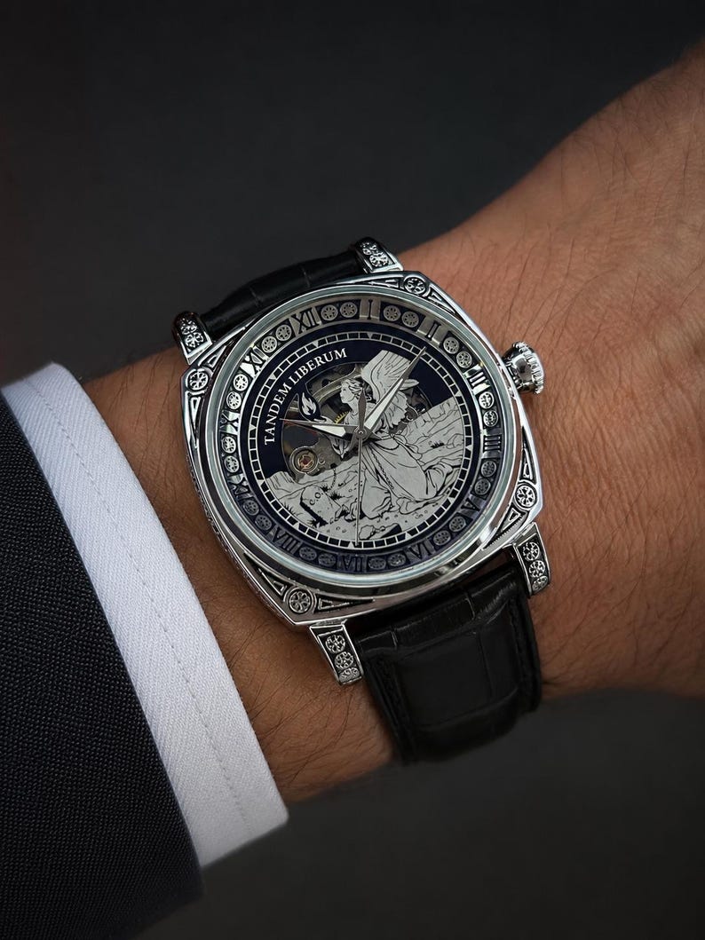Puede incluir: Un reloj de pulsera plateado con correa de cuero negro, que presenta una caja cuadrada con grabados intrincados. La esfera muestra una ilustraci&oacute;n central de un &aacute;ngel, el texto TANDEM LIBERUM y un borde con n&uacute;meros romanos y motivos circulares.