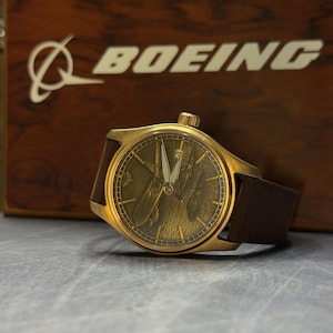 May include: A gold wristwatch with a brown leather strap. The watch face has a detailed design, and the word "BOEING" is visible in the background on a wooden surface.