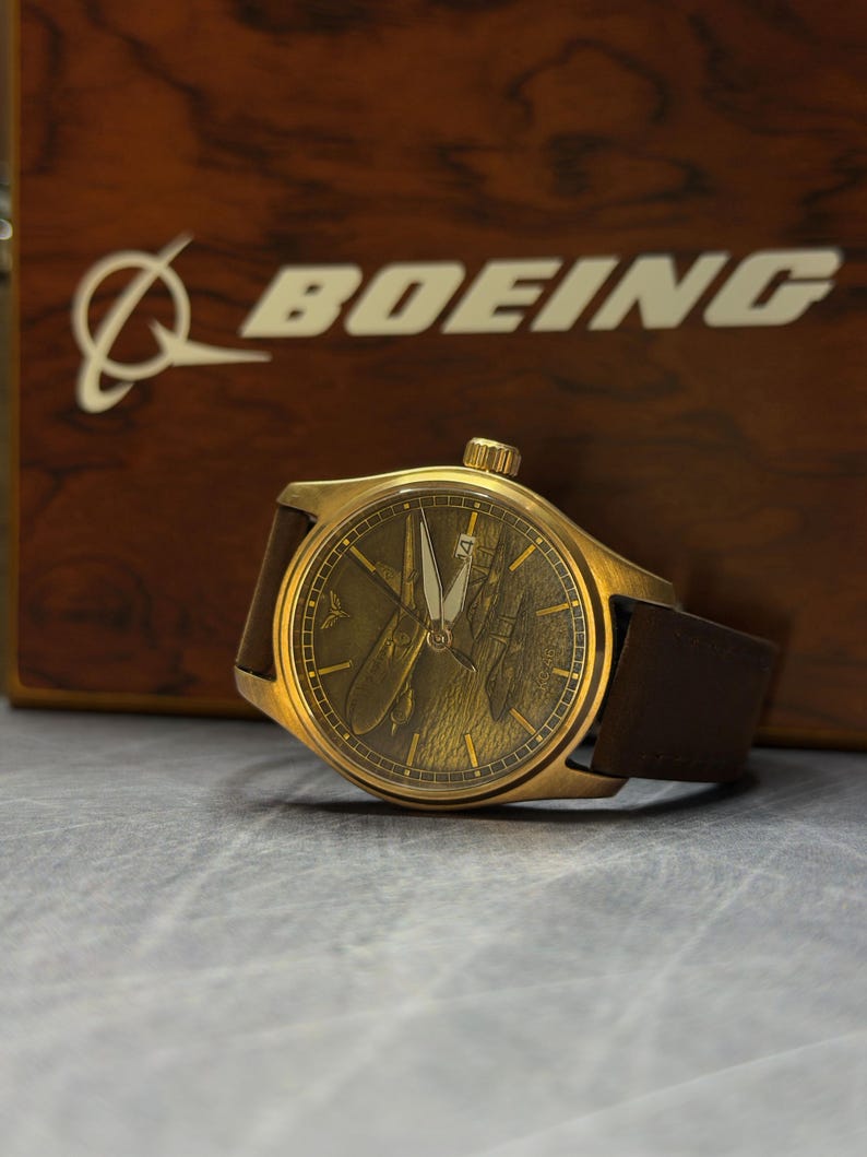May include: A gold wristwatch with a brown leather strap. The watch face has a detailed design, and the word "BOEING" is visible in the background on a wooden surface.