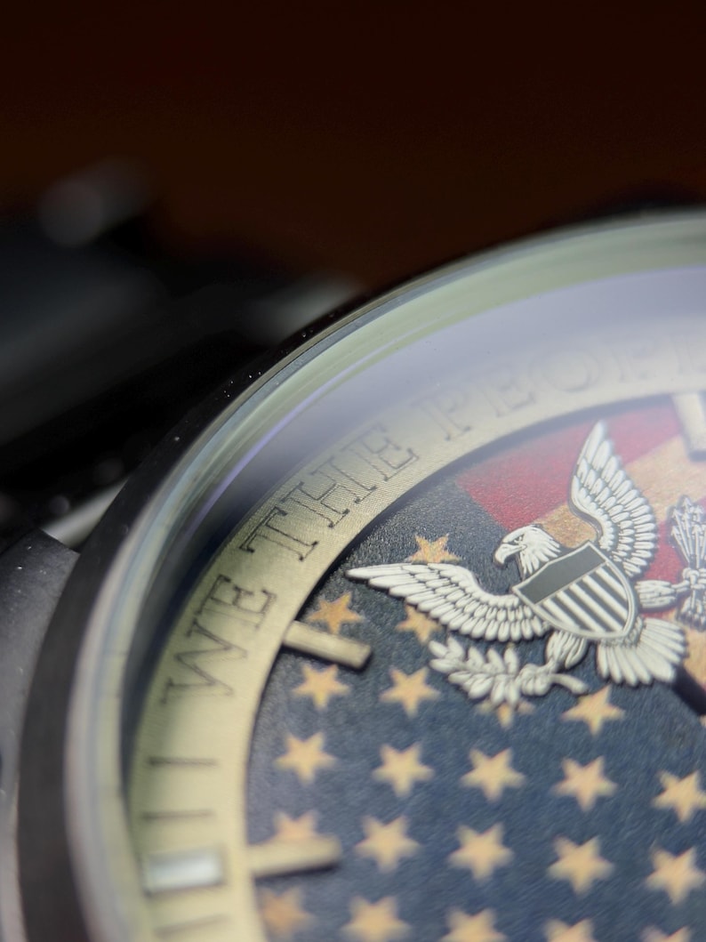 K&ouml;nnte beinhalten: Nahaufnahme eines Zifferblatts mit patriotischem Design. Das Zifferblatt hat einen blauen Hintergrund mit goldenen Sternen, einen rot-wei&szlig; gestreiften Abschnitt und ein detailliertes Adleremblem. Die Worte "WE THE PEOPLE" sind sichtbar.