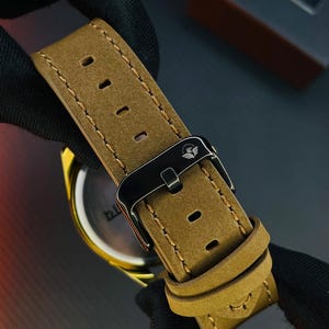 K&ouml;nnte beinhalten: Ein beiges Uhrenarmband aus Wildleder mit hellbraunen Kontrastn&auml;hten und einer schwarzen Metallschnalle, auf deren Oberseite ein kleines wei&szlig;es Adlerlogo eingraviert ist. Das Band hat mehrere rechteckige L&ouml;cher und zwei Schlaufen.
