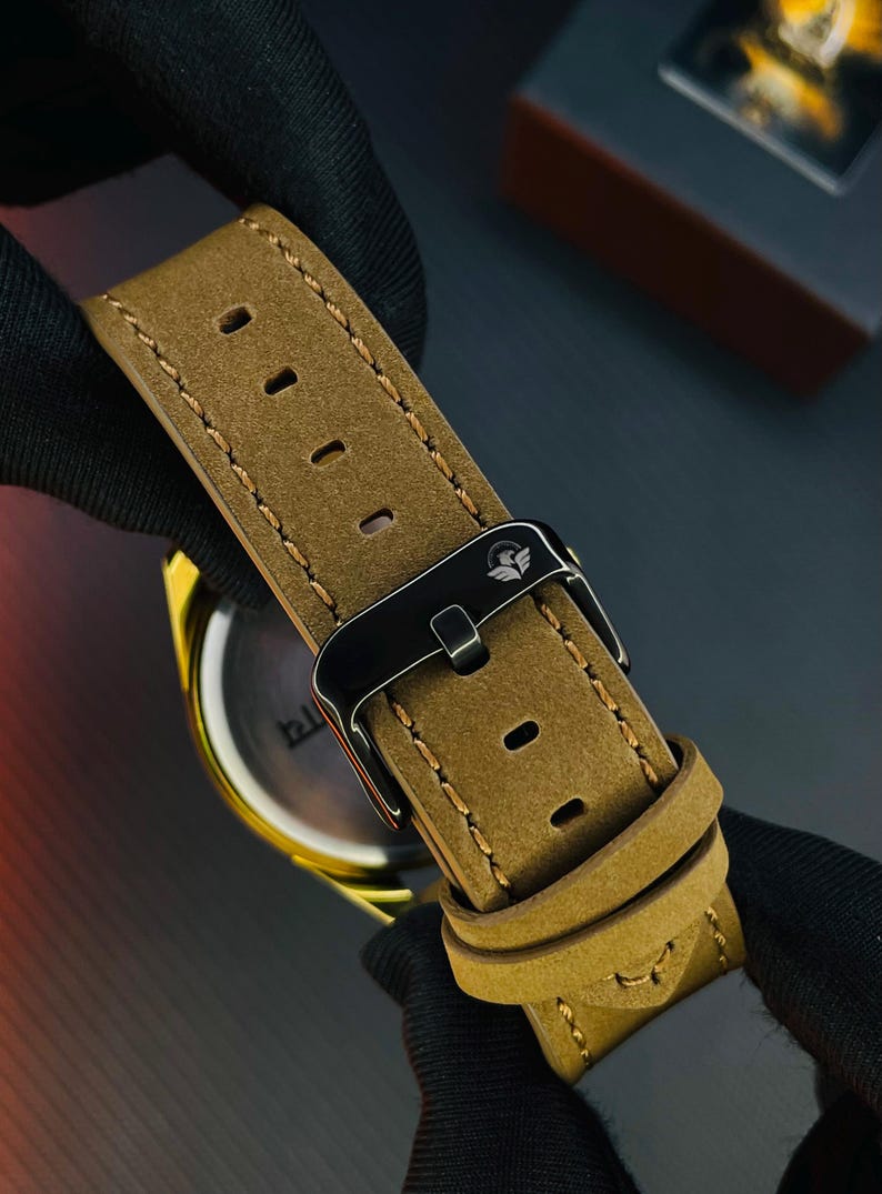 K&ouml;nnte beinhalten: Ein beiges Uhrenarmband aus Wildleder mit hellbraunen Kontrastn&auml;hten und einer schwarzen Metallschnalle, auf deren Oberseite ein kleines wei&szlig;es Adlerlogo eingraviert ist. Das Band hat mehrere rechteckige L&ouml;cher und zwei Schlaufen.