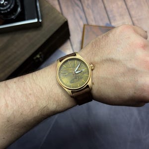 May include: A gold-coloured wristwatch with a brown leather strap is displayed on a wrist. The watch face has a textured, greenish-gold appearance with white hands. The watch is set against a wooden background.