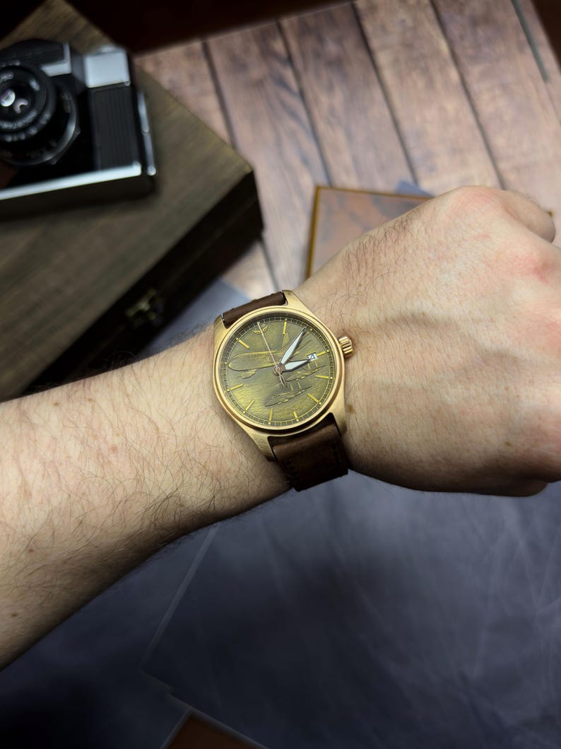 May include: A gold-coloured wristwatch with a brown leather strap is displayed on a wrist. The watch face has a textured, greenish-gold appearance with white hands. The watch is set against a wooden background.