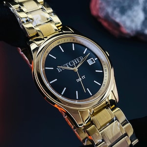 May include: A gold-toned wristwatch with a black face. The watch features the word "BUTCHER" above the center and "DO IT" below. The watch has a date display and a gold link band. The watch is held against a dark background.