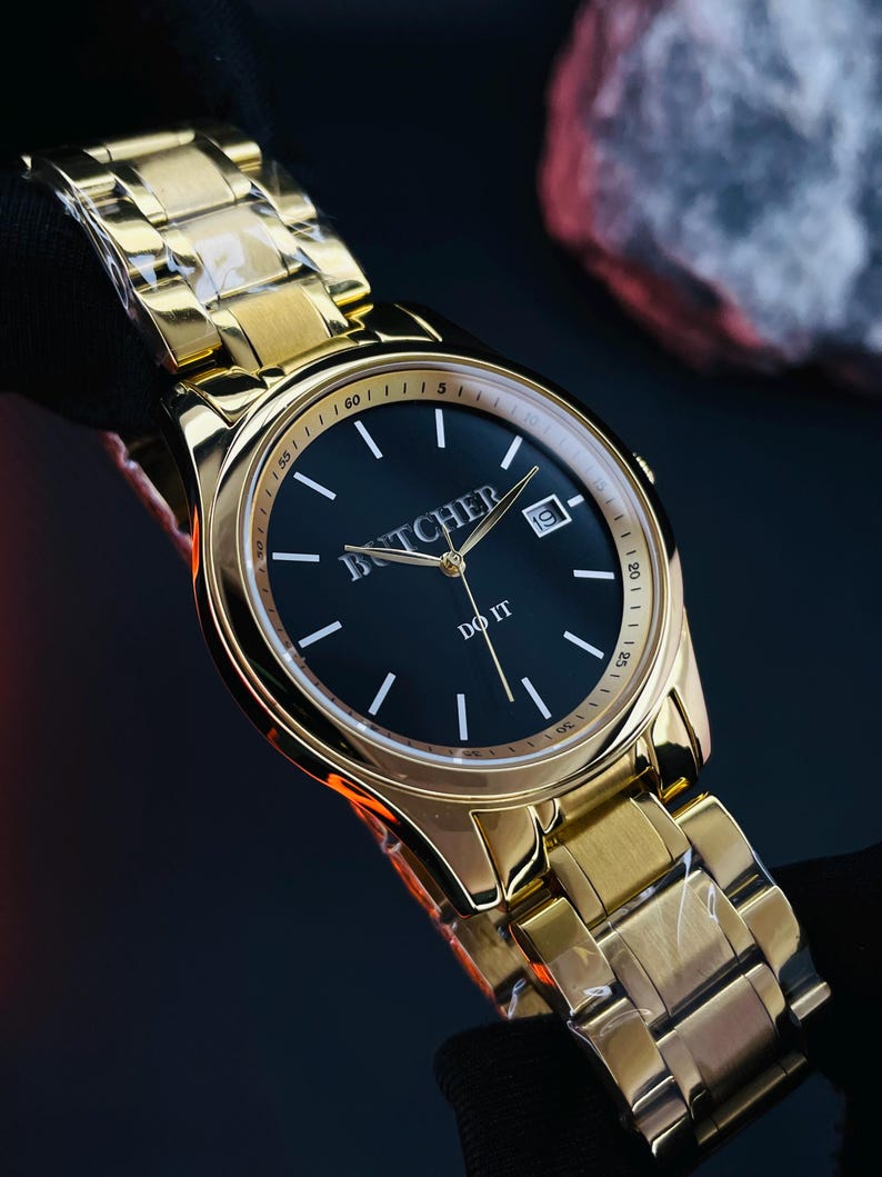 May include: A gold-toned wristwatch with a black face. The watch features the word "BUTCHER" above the center and "DO IT" below. The watch has a date display and a gold link band. The watch is held against a dark background.