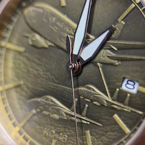 May include: A gold wristwatch with an aviation design. The watch face shows embossed aircraft, including a large plane with "US AIR FORCE" text and fighter jets. The date is visible. The hands are white with gold detailing. The watch has a military theme.