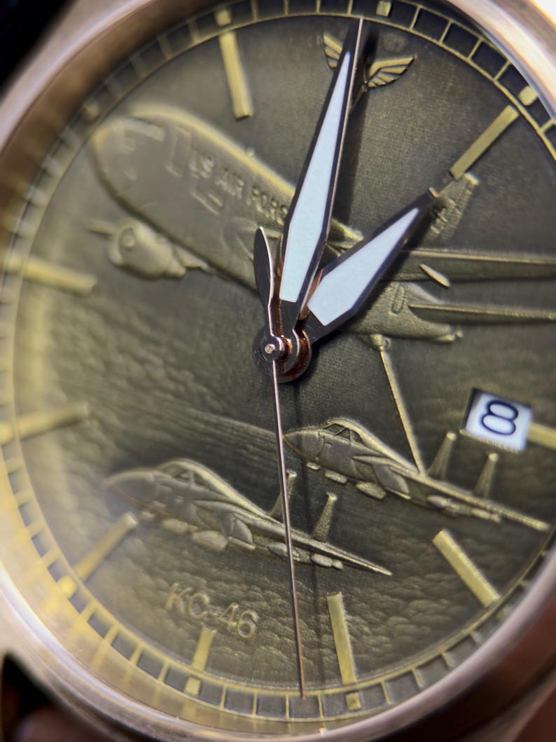 May include: A gold wristwatch with an aviation design. The watch face shows embossed aircraft, including a large plane with "US AIR FORCE" text and fighter jets. The date is visible. The hands are white with gold detailing. The watch has a military theme.