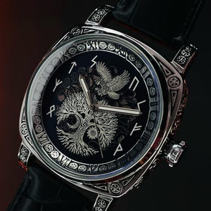 K&ouml;nnte beinhalten: Eine silberfarbene Armbanduhr mit schwarzem Zifferblatt und schwarzem Lederarmband. Das Zifferblatt zeigt ein Baum- und Adlerdesign. Die Uhr hat ein quadratisches Zifferblatt mit aufwendigen Details an der L&uuml;nette und an den Bandans&auml;tzen.