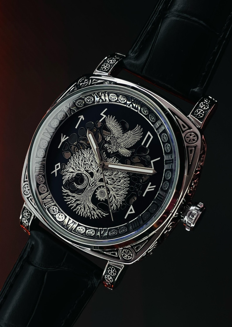 K&ouml;nnte beinhalten: Eine silberfarbene Armbanduhr mit schwarzem Zifferblatt und schwarzem Lederarmband. Das Zifferblatt zeigt ein Baum- und Adlerdesign. Die Uhr hat ein quadratisches Zifferblatt mit aufwendigen Details an der L&uuml;nette und an den Bandans&auml;tzen.