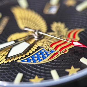 K&ouml;nnte beinhalten: Nahaufnahme eines schwarzen Zifferblatts mit einem goldenen Adleremblem und roten, wei&szlig;en und blauen Akzenten. Das Zifferblatt hat goldene Sterne und leuchtende Zeiger. Die Uhr hat einen strukturierten Hintergrund.