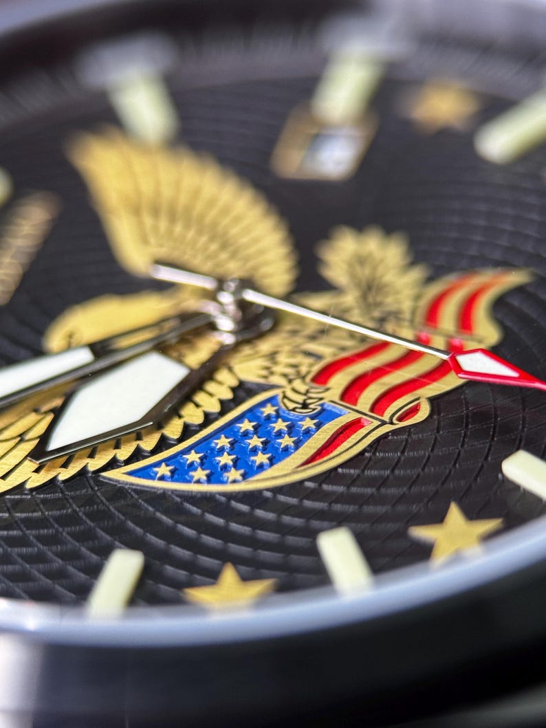 K&ouml;nnte beinhalten: Nahaufnahme eines schwarzen Zifferblatts mit einem goldenen Adleremblem und roten, wei&szlig;en und blauen Akzenten. Das Zifferblatt hat goldene Sterne und leuchtende Zeiger. Die Uhr hat einen strukturierten Hintergrund.