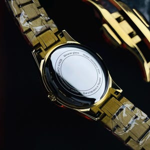 May include: Gold-toned wristwatch with a matching band. The watch face is visible, with the text "Mineral glass" and "Water resistant 30m". The watch band is made of rectangular links. The watch is partially wrapped in clear plastic.