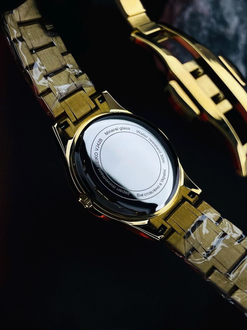 May include: Gold-toned wristwatch with a matching band. The watch face is visible, with the text "Mineral glass" and "Water resistant 30m". The watch band is made of rectangular links. The watch is partially wrapped in clear plastic.