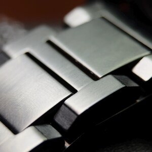 K&ouml;nnte beinhalten: Nahaufnahme eines silbernen Metall-Uhrenarmbands. Das Armband besteht aus rechteckigen Gliedern mit geb&uuml;rsteter Oberfl&auml;che. Die Glieder sind ineinander verschachtelt und bilden eine flexible Struktur. Ein elegantes, modernes Accessoire.
