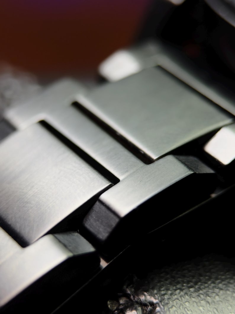 K&ouml;nnte beinhalten: Nahaufnahme eines silbernen Metall-Uhrenarmbands. Das Armband besteht aus rechteckigen Gliedern mit geb&uuml;rsteter Oberfl&auml;che. Die Glieder sind ineinander verschachtelt und bilden eine flexible Struktur. Ein elegantes, modernes Accessoire.
