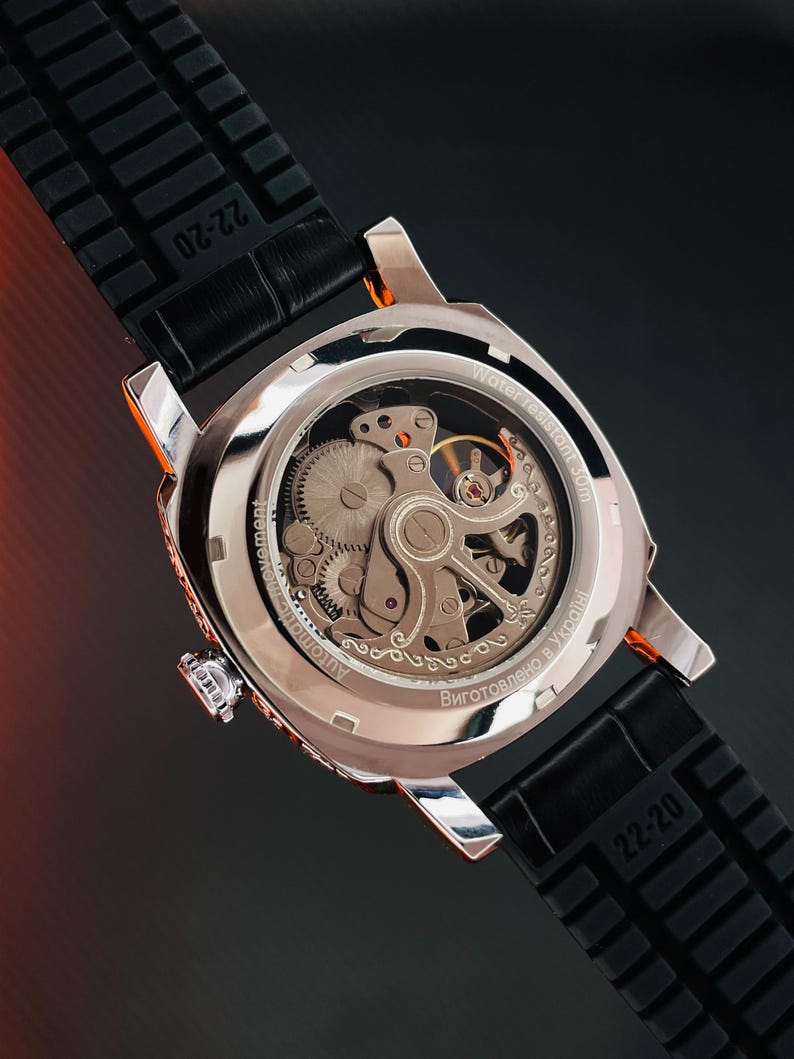 Peut inclure: Gros plan d'une montre automatique argent&eacute;e avec un bracelet en caoutchouc noir. Le dos de la montre r&eacute;v&egrave;le les m&eacute;canismes internes. Le bracelet est marqu&eacute; "22-20". La montre est &eacute;tanche &agrave; 30m.