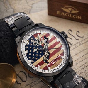 K&ouml;nnte beinhalten: Schwarze Armbanduhr mit patriotischem Design. Das Zifferblatt zeigt die amerikanische Flagge, ein Adleremblem und den Text "1776-2026" und "250th Anniversary Edition". Die Uhr wird mit einer Holzbox pr&auml;sentiert.