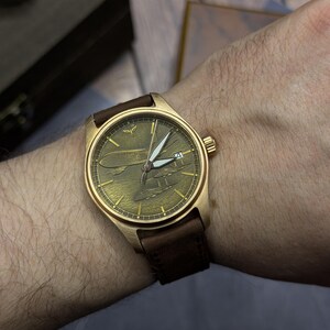 May include: A bronze wristwatch with a brown leather strap. The watch face has an aviation-themed design with white hands and a date display. The watch is on a wrist.