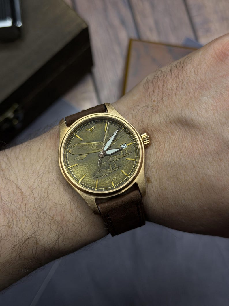 May include: A bronze wristwatch with a brown leather strap. The watch face has an aviation-themed design with white hands and a date display. The watch is on a wrist.