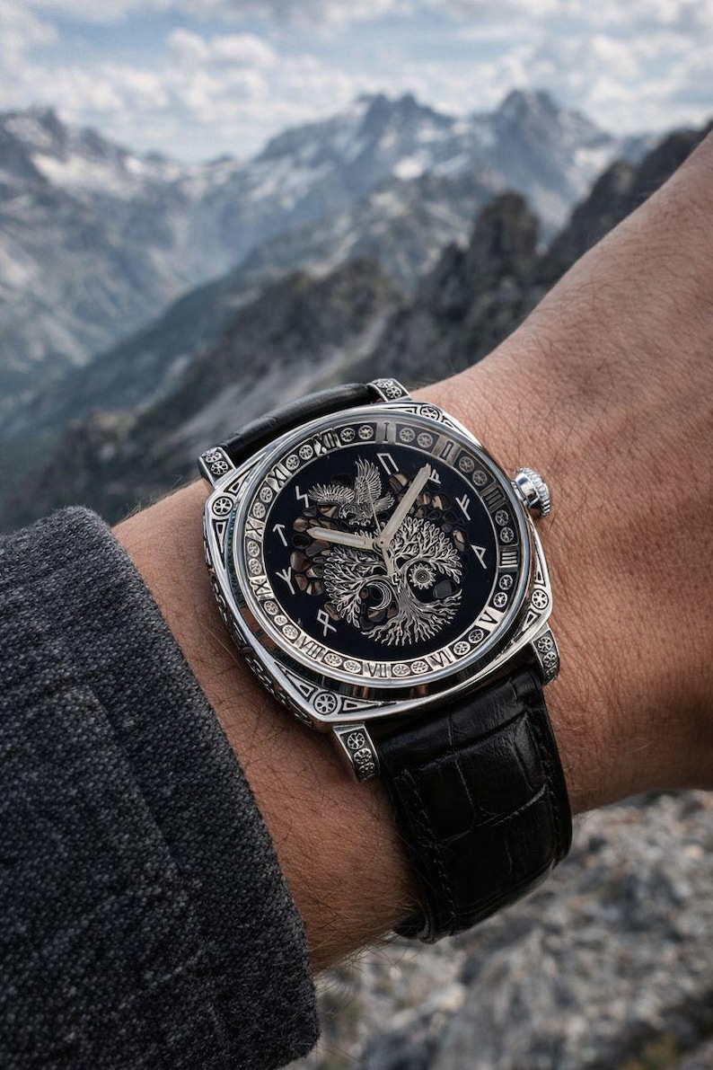 May include: A detailed wristwatch with a black face featuring a tree and bird design, surrounded by silver Roman numerals and symbols. The watch has a silver case and a black leather band. The background shows a mountain range.