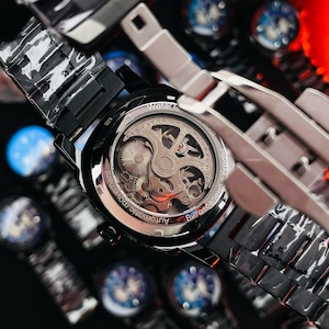 May include: Close-up of a black automatic watch with a transparent back, showcasing the intricate inner workings. The watch has a black band and a silver-colored bezel. The text "Automatic movement" is visible on the watch face. Other watches are visible in the background.