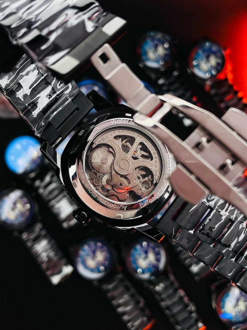 May include: Close-up of a black automatic watch with a transparent back, showcasing the intricate inner workings. The watch has a black band and a silver-colored bezel. The text "Automatic movement" is visible on the watch face. Other watches are visible in the background.