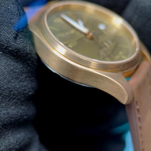 May include: A gold-coloured wristwatch with a brown leather strap. The watch face is a muted green with gold hands and markers. The watch is shown against a dark background, emphasising its metallic finish and design.