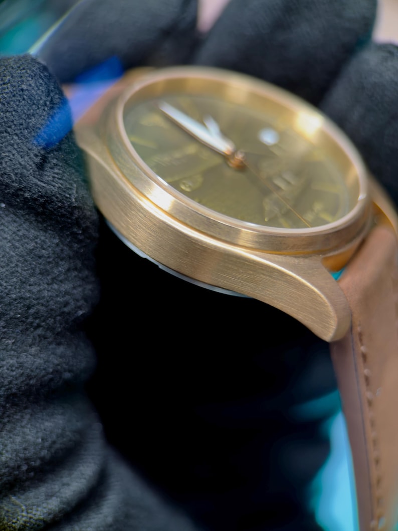 May include: A gold-coloured wristwatch with a brown leather strap. The watch face is a muted green with gold hands and markers. The watch is shown against a dark background, emphasising its metallic finish and design.